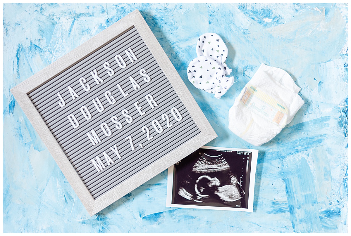 Photograph of newborn baby details on a blue mat including a letter board, which reads: Jackson Douglas Mosser, May 7, 2020, with hand covers, ultrasound photo and premie size diaper.